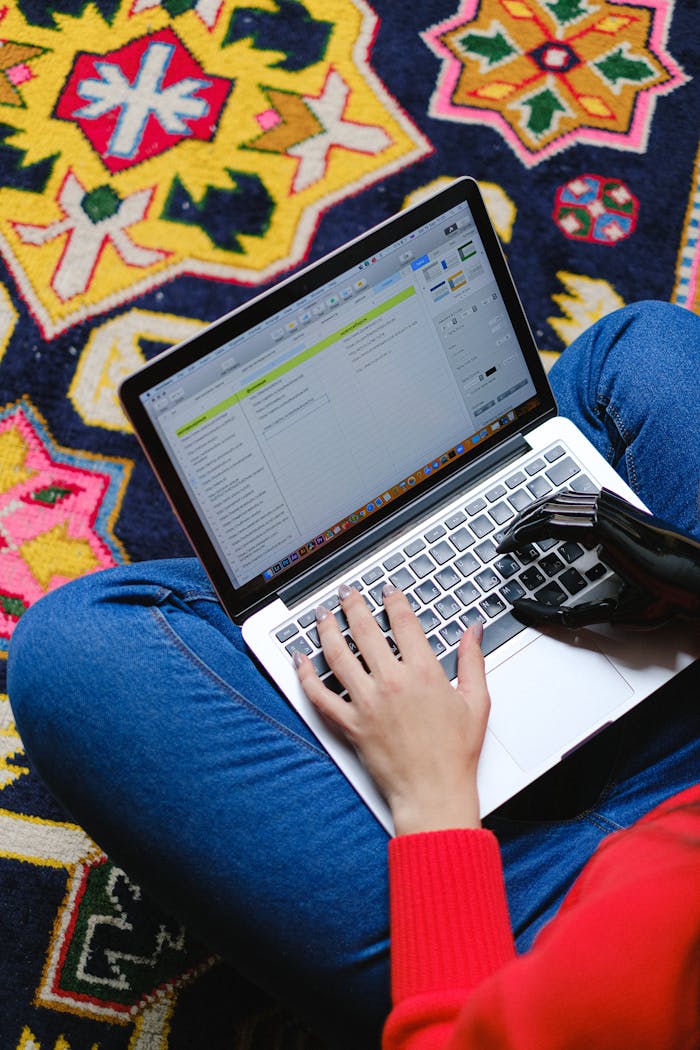 A person with a prosthetic hand typing on a laptop while sitting on a colorful patterned rug.